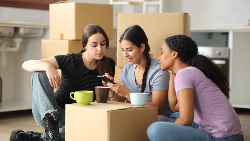 View of three young females looking at a mobile phone amongst moving boxes